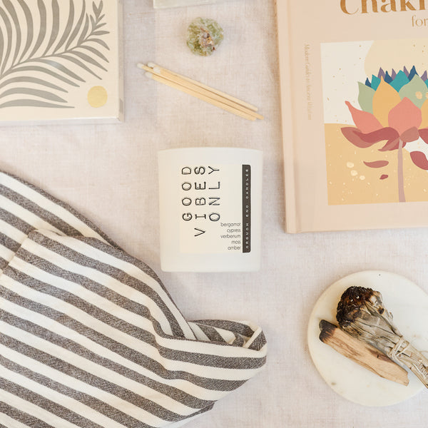 Candle labeled 'Good Vibes Only' on a surface with books and a striped towel.