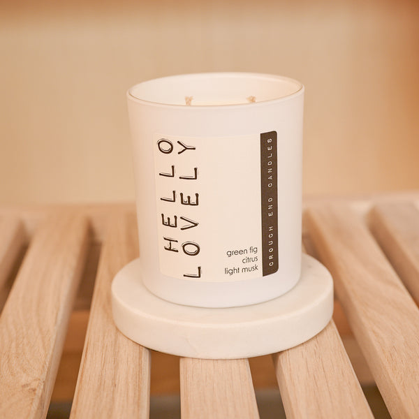White candle labeled 'HELLO LOVELY' on a wooden stand with a warm glow.