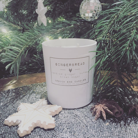 A white candle with 'GINGERBREAD' text on it, placed on a table with decorative cookies, a star-shaped cookie, and a pine tree in the background.